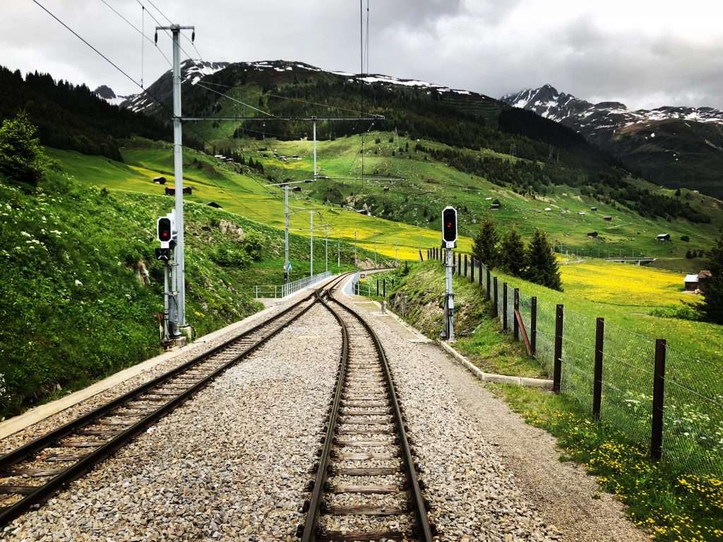 photo of a railroad track in switzerland with mountains in the background and yellow flowers on the green grass